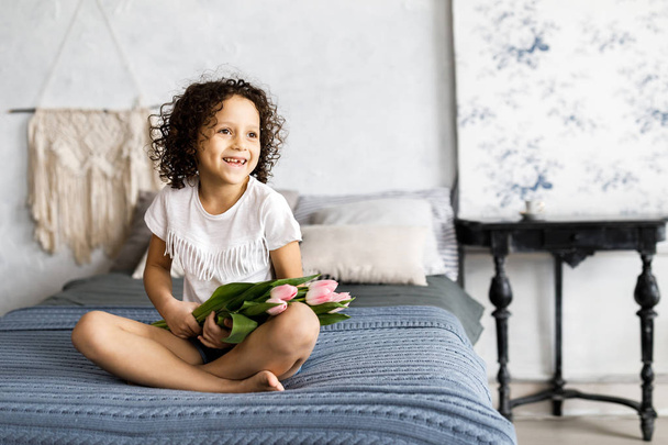 Маленькая красивая кудрявая девочка, сидящая на кровати с тюльпанами на руках.
 - Фото, изображение