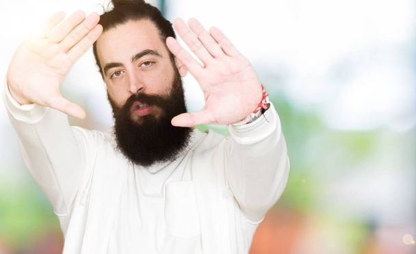 Homem jovem hipster com cabelos longos e barba vestindo camisola esportiva Sorrindo fazendo quadro usando as mãos palmas e dedos, perspectiva da câmera
 - Foto, Imagem