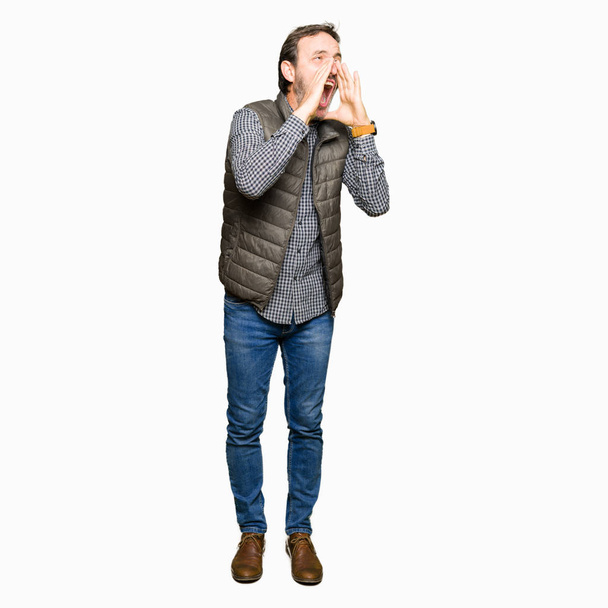 Knappe man van middelbare leeftijd dragen winter vest schreeuwen hardop vertoornd overhandigt mond - Foto, afbeelding