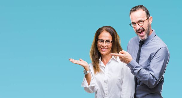 Middle age hispanic couple in love wearing glasses over isolated background amazed and smiling to the camera while presenting with hand and pointing with finger. - Photo, Image