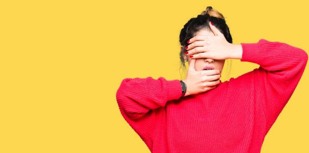 Young beautiful woman wearing red sweater and bun Covering eyes and mouth with hands, surprised and shocked. Hiding emotion - Фото, зображення