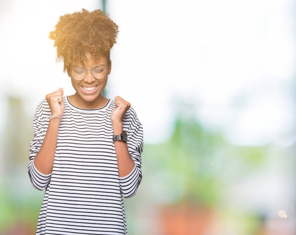 Красивая молодая африканская американка в очках на изолированном фоне, взволнованная успехом с поднятыми руками, с улыбкой отмечающая победу. Концепция победителя
. - Фото, изображение