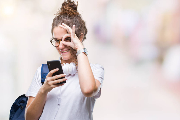 Dziewczyna młoda brunetka student szczęśliwy twarz uśmiechający się robi ok znak ręką na oko patrząc przez palce na sobie plecak, słuchawki i smartphone na białym tle - Zdjęcie, obraz
