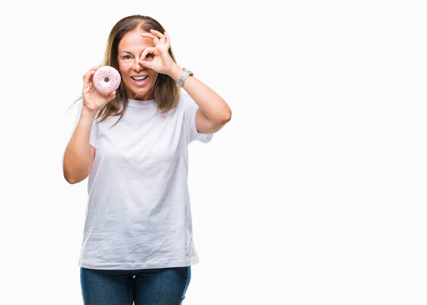 Orta yaş İspanyol kadın mutlu yüz el parmaklarının arasından seyir göz üzerinde ok işaretiyle yapıyor gülümseyen izole arka plan üzerinde pembe çörek yeme - Fotoğraf, Görsel