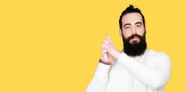 Young hipster man with long hair and beard wearing sporty sweatshirt Holding symbolic gun with hand gesture, playing killing shooting weapons, angry face - Foto, Imagem
