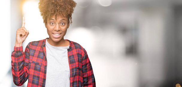 Başarılı fikirle izole arka plan işaret parmağı üzerinde gözlük takan güzel genç Afro-Amerikan kadın. Çıktıktan ve mutlu. Bir numara. - Fotoğraf, Görsel