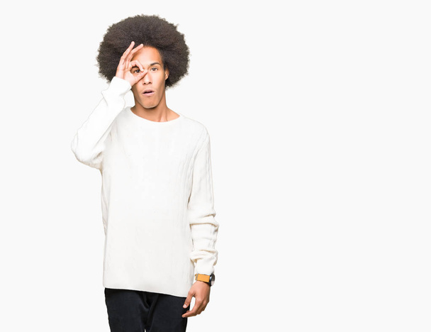 Young african american man with afro hair wearing winter sweater doing ok gesture shocked with surprised face, eye looking through fingers. Unbelieving expression. - Foto, Bild