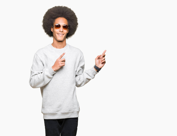 Giovane uomo afro-americano con i capelli afro indossare occhiali da sole sorridente e guardando la fotocamera che punta con due mani e le dita di lato
. - Foto, immagini