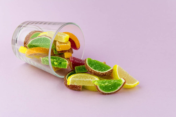 Multi-colored marmalade in the form of citrus slices in a transparent glass, which lies on a pink background - Фото, изображение
