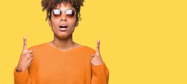 Mooie jonge african american vrouw dragen van een zonnebril op geïsoleerde achtergrond verbaasd en verrast opzoeken en wijzen met vingers en opgeheven armen. - Foto, afbeelding