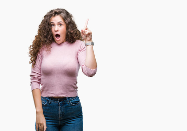 Beautiful brunette curly hair young girl wearing pink sweater over isolated background pointing finger up with successful idea. Exited and happy. Number one. - 写真・画像