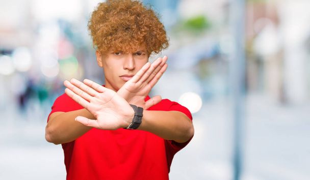 Jeune homme beau aux cheveux afro portant un t-shirt rouge Expression de rejet croisant les bras et les paumes faisant signe négatif, visage en colère
 - Photo, image