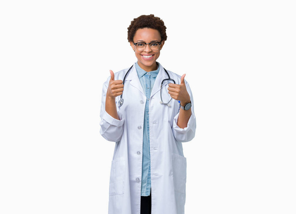 手で、肯定的なジェスチャーを行う分離背景成功記号の上医療のコートを着ている若いアフリカ系アメリカ人医師女性の親指を笑顔と幸せ。勝者ジェスチャー晴れやかな表情でカメラを見てください。. - 写真・画像