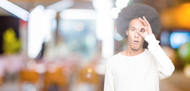 Americký mladík s afro vlasy nosit zimní svetr dělá ok gesto šokoval s tváří překvapeně, oko dívá skrze prsty. Nevěřící výraz. - Fotografie, Obrázek
