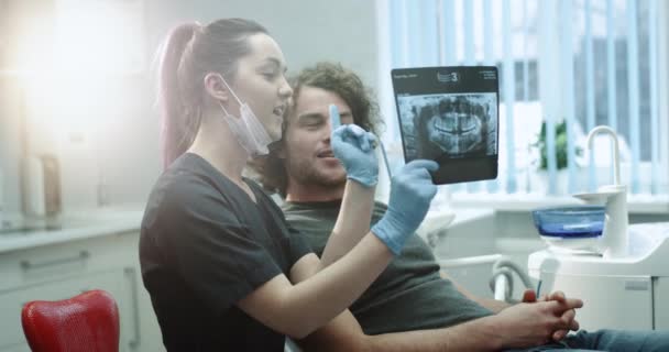 Young dentist woman with blue gloves and mask explain to her patient his result of teeth X-ray , showing to the man his problem , they have a nice conversation , smiling faces in a light dentist room - Πλάνα, βίντεο