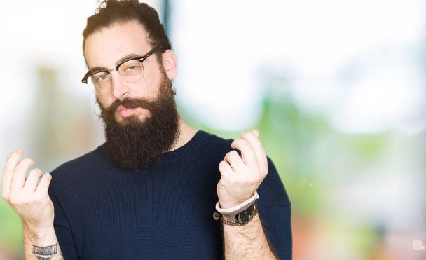 Young hipster man with long hair and beard wearing glasses Doing money gesture with hand, asking for salary payment, millionaire business - Foto, Imagem