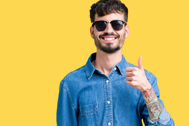 Jonge knappe man dragen van een zonnebril op geïsoleerde achtergrond gelukkig duimschroef opwaarts gebaar met de hand te doen. Goedkeuring van expressie kijken naar de camera met tonen van succes. - Foto, afbeelding