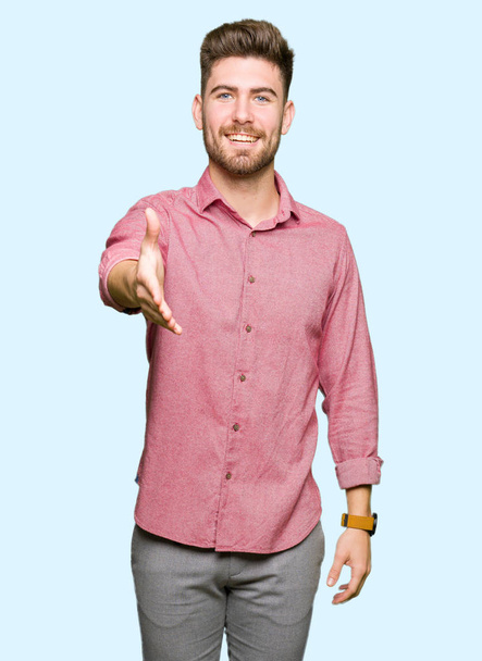 Młody przystojny business casual człowiek uśmiechający się oferujący przyjazny uścisk dłoni jako pozdrowienie i przyjazna. Udanego biznesu. - Zdjęcie, obraz