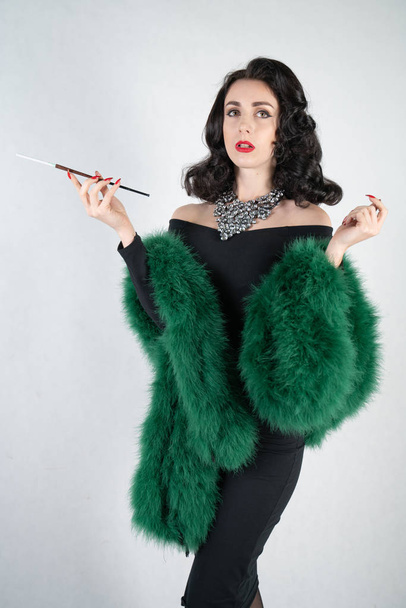 charming graceful pin up girl in a black evening dress and a green fluffy boa in her hands stands and smokes a cigarette with a mouthpiece on a white solid Studio background - Фото, изображение