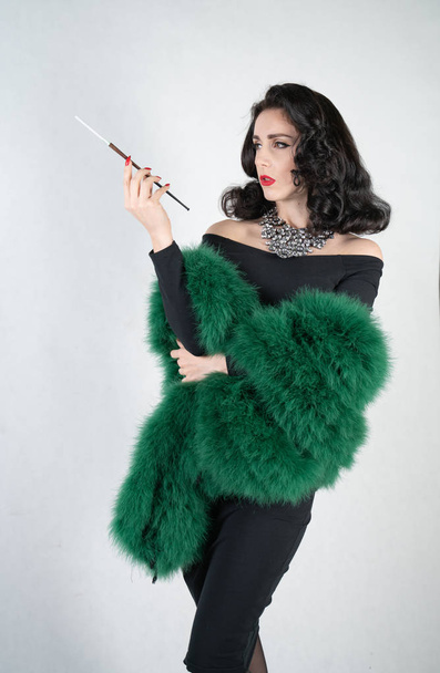 charming graceful pin up girl in a black evening dress and a green fluffy boa in her hands stands and smokes a cigarette with a mouthpiece on a white solid Studio background - Foto, Bild
