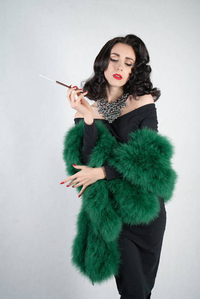 charming graceful pin up girl in a black evening dress and a green fluffy boa in her hands stands and smokes a cigarette with a mouthpiece on a white solid Studio background - Φωτογραφία, εικόνα