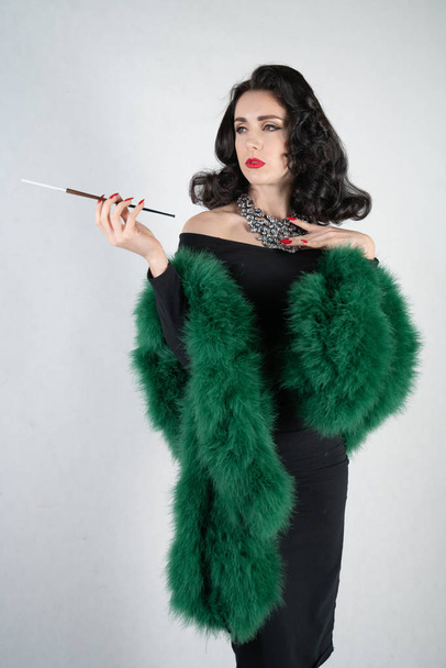 charming graceful pin up girl in a black evening dress and a green fluffy boa in her hands stands and smokes a cigarette with a mouthpiece on a white solid Studio background - 写真・画像