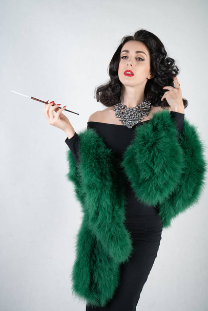 charming graceful pin up girl in a black evening dress and a green fluffy boa in her hands stands and smokes a cigarette with a mouthpiece on a white solid Studio background - Photo, Image