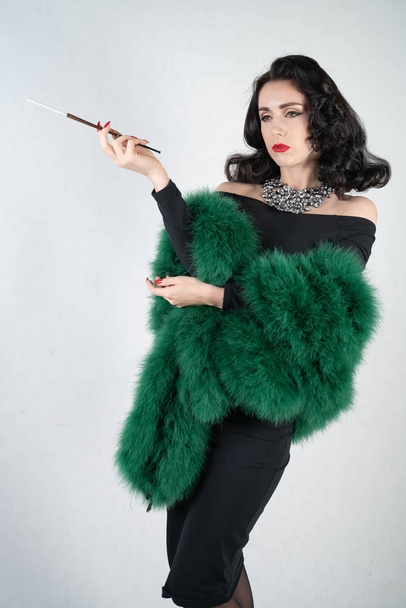 charming graceful pin up girl in a black evening dress and a green fluffy boa in her hands stands and smokes a cigarette with a mouthpiece on a white solid Studio background - Fotografie, Obrázek