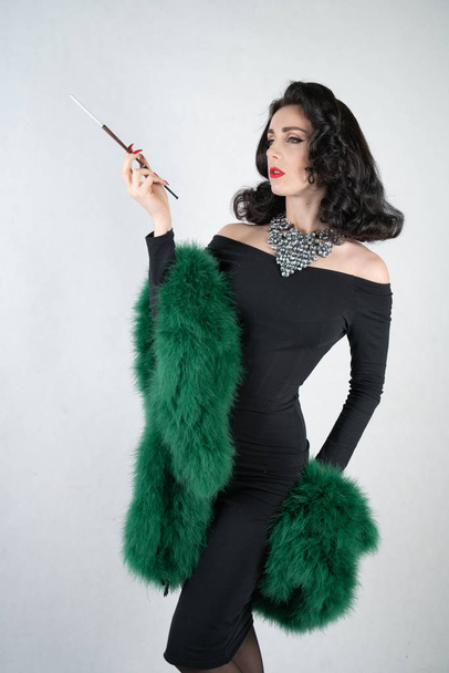 γοητευτικό χαριτωμένη pin up κορίτσι σε ένα μαύρο φόρεμα βράδυ και ένα πράσινο χνουδωτό boa στα χέρια της στέκεται και καπνίζει ένα τσιγάρο με επιστόμιο σε λευκό στερεό φόντο στούντιο - Φωτογραφία, εικόνα