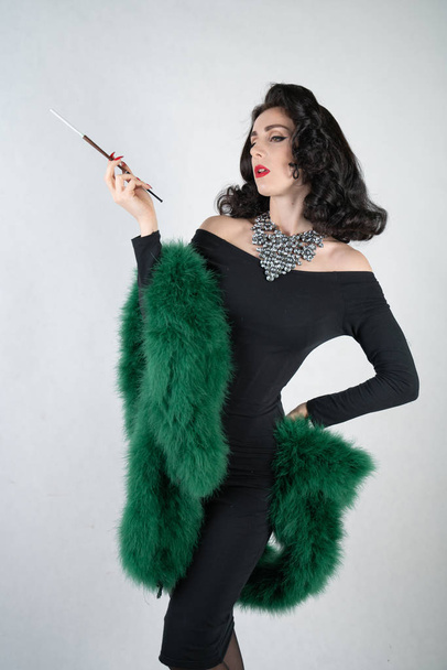 charmante sierlijke pin up meisje in een zwarte avond jurk en een groene pluizige boa in haar handen staat en rookt een sigaret met een mondstuk op een witte achtergrond voor solide Studio - Foto, afbeelding