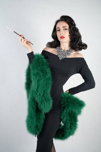 charming graceful pin up girl in a black evening dress and a green fluffy boa in her hands stands and smokes a cigarette with a mouthpiece on a white solid Studio background - Photo, Image