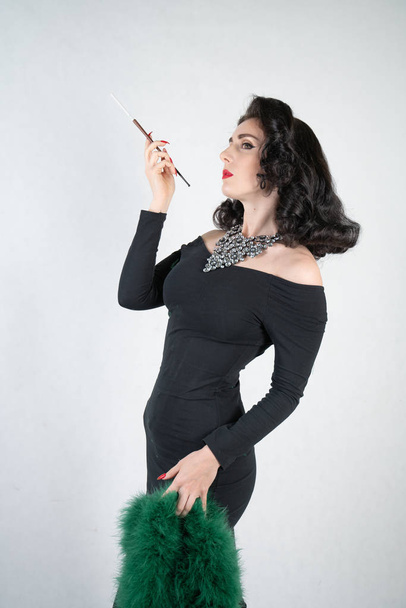 bájos, kecses pin-up lány egy fekete estélyi ruha és egy zöld bolyhos boa a kezében áll, és egy cigarettát, fehér alapon szilárd Studio csutorával - Fotó, kép