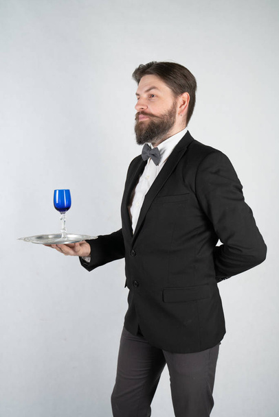 Кавказский слуга с бородой в официальном деловом костюме стоит со стальным подносом в руке и бокалом вина на белом твердом фоне в Студии
 - Фото, изображение