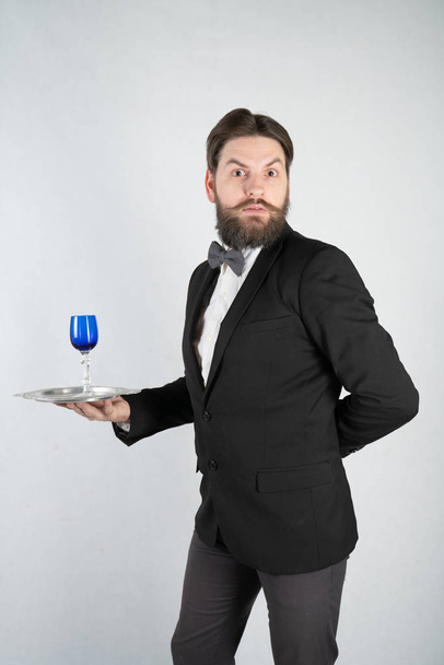 caucasian servant with a beard in a formal business suit stands with a steel tray in his hand and a glass of wine on a white solid background in the Studio - Foto, immagini