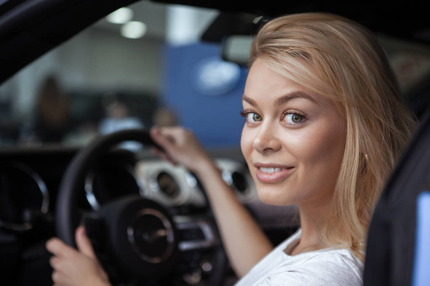 Close up of beautiful blond haired woman smiling over her shoulder to the camera, sitting behind the steering wheel of a new auto. Gorgeous cheerful female enjoying buying new car. Dealership concept - Фото, зображення