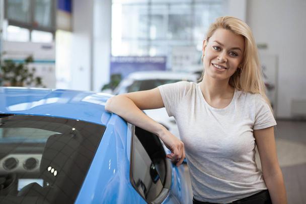 Young happy beautiful woman smiling joyfully after buying new car at the dealership. Charming female driver leaning on a new automobile. Modern car discount offer. Sales, consumerism, retail concept - Fotografie, Obrázek