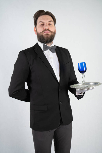 Λευκός υπηρέτης με γενειάδα σε επίσημο επιχειρηματικό κοστούμι στέκεται με ένα δίσκο χάλυβα στο χέρι του και ένα ποτήρι κρασί σε ένα λευκό στερεό φόντο στο στούντιο - Φωτογραφία, εικόνα