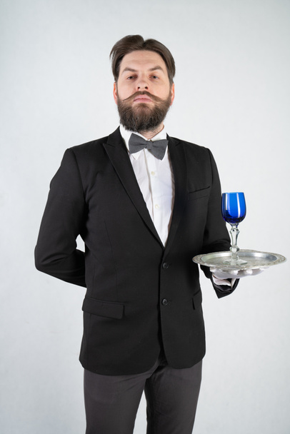 Kaukasischer Diener mit Bart im formellen Businessanzug steht mit einem Stahltablett in der Hand und einem Glas Wein auf weißem Hintergrund im Atelier - Foto, Bild