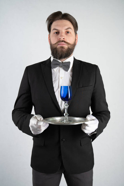 servitore caucasico con la barba in un abito da lavoro formale sta con un vassoio di acciaio in mano e un bicchiere di vino su uno sfondo solido bianco nello Studio
 - Foto, immagini