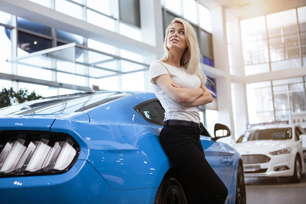Low angle shot of a gorgeous blonde leaning on her beautiful new car, posing at the dealership salon. Attractive woman buying muscle car. Happy female looking away dreamily after buying new auto - Fotografie, Obrázek
