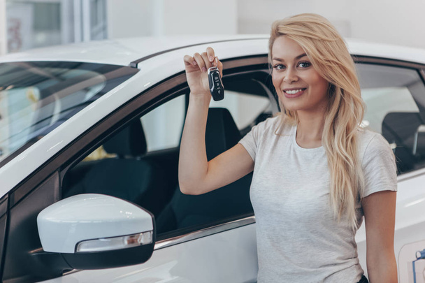 Young happy female driver holding car key, posing near her new automobile at dealership salon, copy space. Charming woman smiling to the camera after buying new auto, copy space. Owner, driver concept - Фото, изображение
