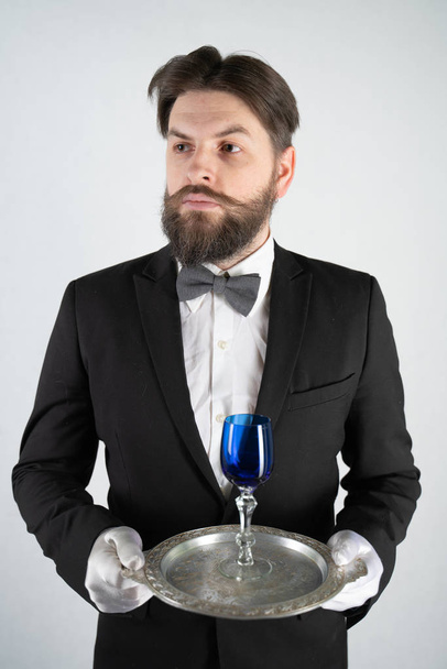 serviteur caucasien avec une barbe dans un costume d'affaires formelle se tient avec un plateau en acier à la main et un verre de vin sur un fond blanc solide dans le studio
 - Photo, image