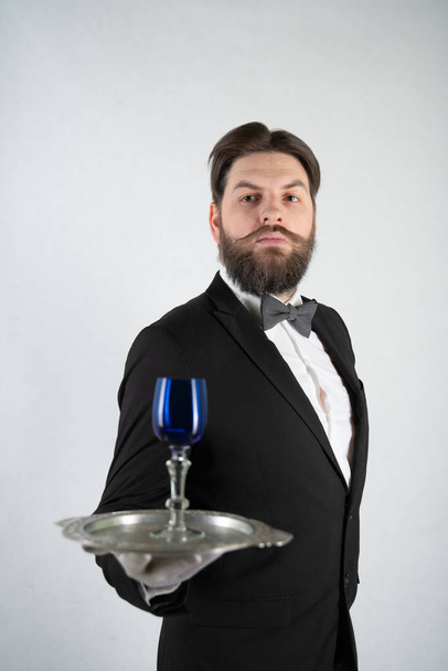 caucasian servant with a beard in a formal business suit stands with a steel tray in his hand and a glass of wine on a white solid background in the Studio - Photo, Image