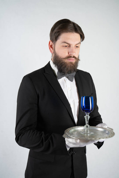 serviteur caucasien avec une barbe dans un costume d'affaires formelle se tient avec un plateau en acier à la main et un verre de vin sur un fond blanc solide dans le studio
 - Photo, image
