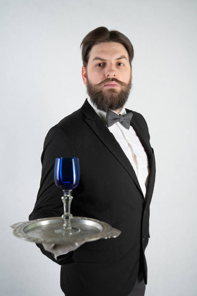 caucasian servant with a beard in a formal business suit stands with a steel tray in his hand and a glass of wine on a white solid background in the Studio - Foto, Imagem
