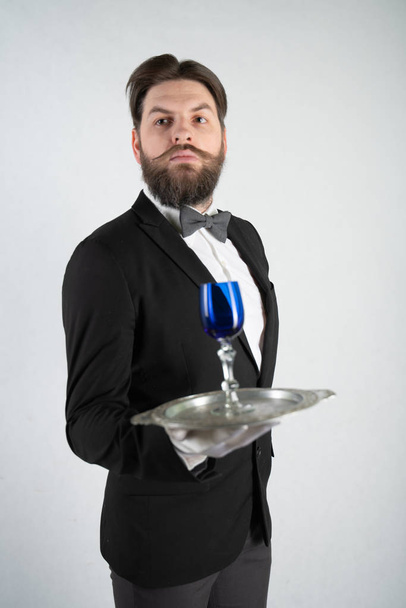 kaukázusi szolga a szakálla a hivatalos üzleti öltöny áll egy acél tálcát a kezében, és egy pohár bort egy fehér, szilárd háttér a stúdióban - Fotó, kép
