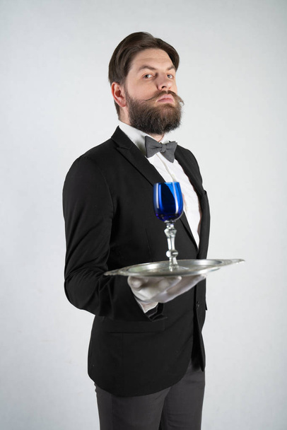 Λευκός υπηρέτης με γενειάδα σε επίσημο επιχειρηματικό κοστούμι στέκεται με ένα δίσκο χάλυβα στο χέρι του και ένα ποτήρι κρασί σε ένα λευκό στερεό φόντο στο στούντιο - Φωτογραφία, εικόνα