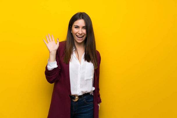 Mujer joven sobre pared amarilla saludando con la mano con expresión feliz
 - Foto, imagen