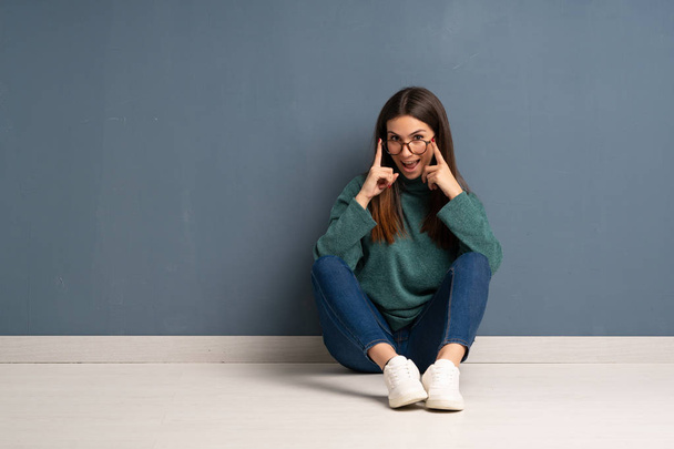 Young woman sitting on the floor with glasses and surprised - Foto, Imagem