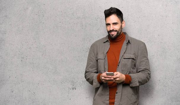 Handsome man with beard sending a message with the mobile over textured wall - Photo, Image